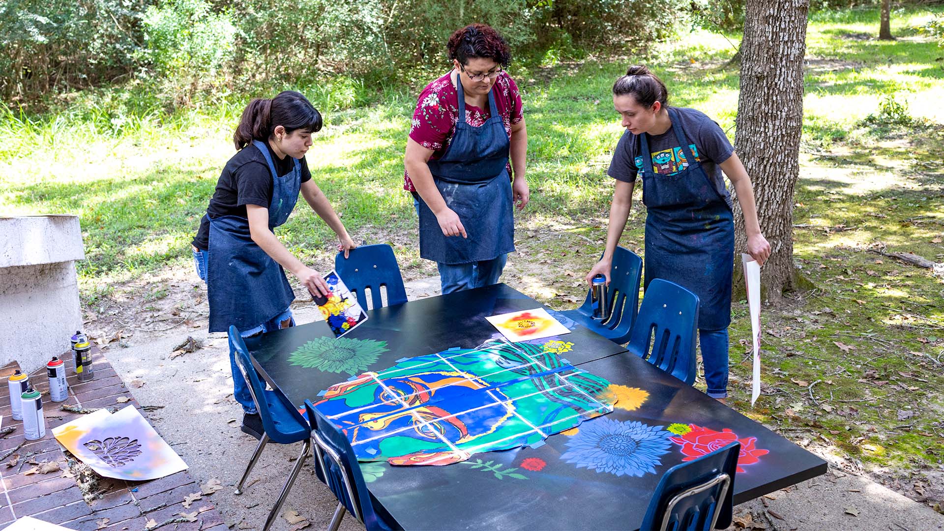 Three students in aprons gathered around a Frida Kahlo mural, set up on a low table outdoors